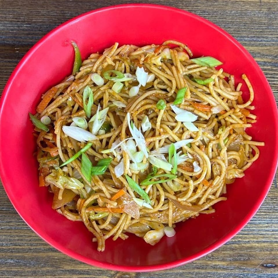 Stir-fried noodles with mixed vegetables and sliced scallions served in a bright red bowl on a wooden table.