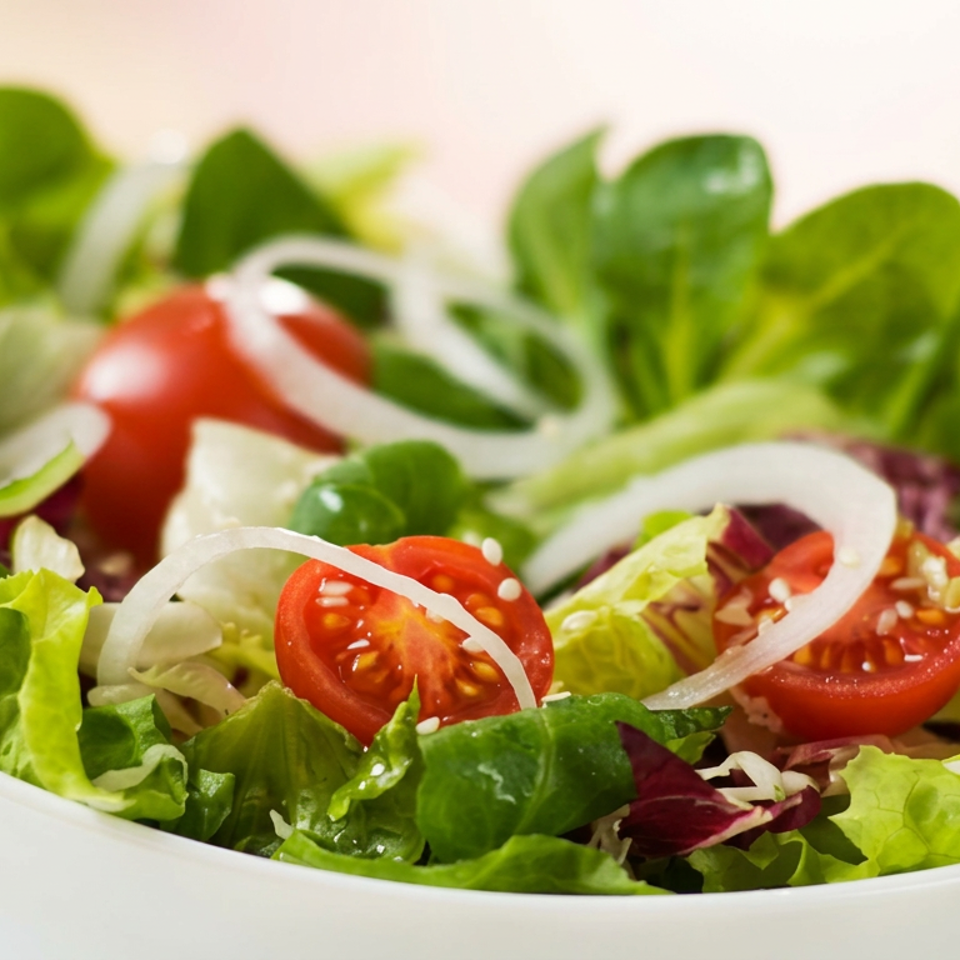 Fresh mixed green salad with cherry tomatoes, sliced onions, cucumber, and sesame seeds in a white bowl.